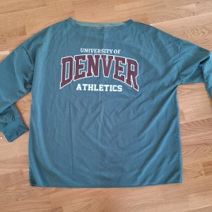 Grön långärmad Denver college-tröja oversized XL - College-tröja i grön färg med tryck 'University of Denver Athletics' i vitt och vinrött framtill. Långärmad modell med rund hals och ribbade muddar. Perfekt för en avslappnad och sportig stil.