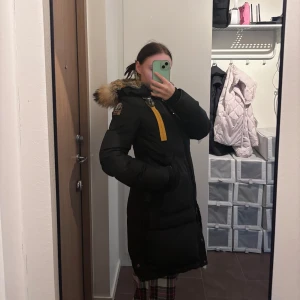 Svart lång dunjacka Parajumpers XS - Lång svart dunjacka från Parajumpers med gul detalj vid dragkedjan och stor huva med äkta päls. Jackan har broderad patch på ärmen och robusta metalldetaljer. Perfekt för kalla dagar och riktigt snygg siluett. Har haft den i flera år. Vid snabb affär kan sänka priset. 