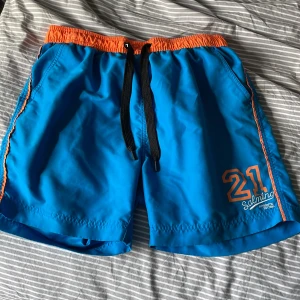 Blå badshorts från Salming - Snygga blå badshorts från Salming med orange midjeresår och detaljer. Svart snörning i midjan och tryckt nummer 21 framtill. Perfekta för stranden eller poolen, med sportig vibe och bekväm passform.