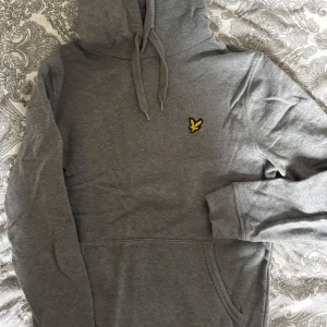 Grå hoodie från Lyle & Scott - Säljer en grå hoodie från Lyle & Scott med klassisk gul logga på bröstet. Tröjan har huva med snörning, magficka och långa ärmar med ribbade muddar. Perfekt för chill dagar eller när du vill ha en enkel och stilren look. Använd ett fåtal gånger. Passar både kille och tjej.