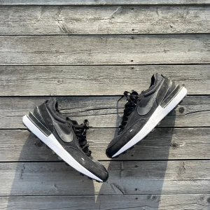 Svarta Nike sneakers med vit sula - Svarta Nike sneakers med klassisk Swoosh-logga på sidan. Ovandelen är i textil och mocka, med svarta skosnören och en vit, chunky sula. Snygg och sportig design som passar perfekt till streetwear. Skorna har rund tå och platt sula för en bekväm känsla.