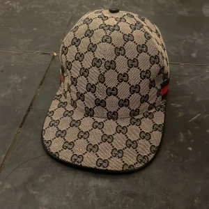 Gucci keps med GG-mönster - Snygg keps från Gucci med klassiskt GG-mönster i svart på beige bakgrund. Kepsen har en böjd skärm och en röd-blå rand på sidan. Tillverkad i bomull med insida i svart tyg. Perfekt för dig som vill ha en lyxig och trendig accessoar.