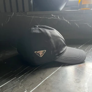 Svart keps från Prada - Snygg svart keps från Prada med klassisk böjd skärm och ikonisk metalltriangel med logga på sidan. Tillverkad i slitstark polyester med justerbar rem baktill för perfekt passform. Perfekt accessoar för dig som vill ha en clean och lyxig look.