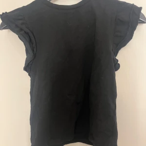 Svart volangärmad topp i bomull - En svart topp med volangdetaljer på ärmarna och rund halsringning. Modellen är ärmlös med en loose passform och tillverkad i mjuk bomull. Perfekt för en chill och stilren look.