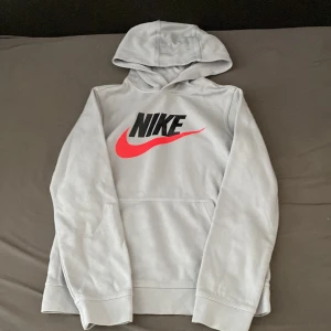 Grå Nike hoodie med röd swoosh - Snygg grå hoodie från Nike med stor svart logga och röd swoosh på bröstet. Klassisk känguruficka och huva. Perfekt för dig som gillar sportig och avslappnad stil. Standard fit och mjukt material.