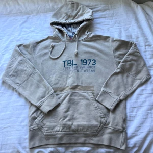 Beige hoodie - Timberland   - Säljer en beige hoodie från Timberland med blått tryck 'TBL 1973' på bröstet. Tröjan har huva med snörning, känguruficka och rutigt foder i luvan. Perfekt för dig som gillar avslappnad och klassisk streetstil. Strl - M