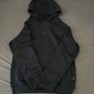 Svart Nike Air hoodie med huva - Svart hoodie från Nike med Air-logga på bröstet och klassisk swoosh. Tröjan har huva, långa ärmar och ribbade muddar. Perfekt för dig som gillar sportig och avslappnad stil. Materialet är mjukt och skönt, troligtvis bomull och polyester.