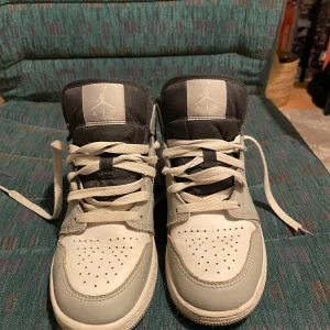 Nike Air Jordan 1 Mid grå/vit - Säljer ett par Nike Air Jordan 1 Mid i grått och vitt med svarta detaljer och klassisk swoosh på sidan. Skorna har snörning, perforerad tå och är tillverkade i skinn och syntet. Perfekta för dig som gillar streetstyle och vill ha en ikonisk sneaker i garderoben. Dessa skor är knappt andvända och kostar runt 3000kr nya! Priset är inte hugget i sten Kan gå ner litegrann.