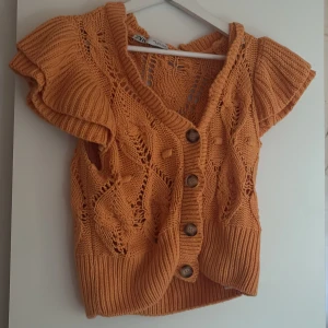 Stickad orange topp med knappar från ZARA - Superfin orange stickad topp från ZARA med korta volangärmar och stora träknappar framtill. Toppen har ett öppet, mönstrat stickat material och bred ribbad kant nedtill. Perfekt för dig som vill ha en färgglad och unik look.