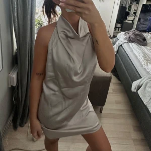 Champagnefärgad satin klänning - Snygg kort klänning från Object i champange/silvergrå färg i satin med halterneck och draperad hals. Klänningen är ärmlös och har en slät, glansig yta som ger en lyxig vibe. Knäpps med två knappar vid nacken. Endast använd en gång och är som ny! 