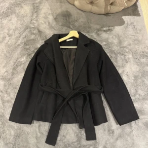 Svart kappa med bälte och krage - Stilren svart kappa med bred krage och knytskärp i midjan. Jackan har en klassisk look med raka ärmar och är helfodrad för extra komfort. Perfekt för dig som gillar en clean och tidlös stil.