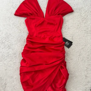 Röd satin klänning från Dressmezee - Supersnygg röd kort klänning från Dressmezee i glansigt satinmaterial. Klänningen har djup v-ringning, draperad design och markerad midja. Perfekt för dig som vill sticka ut med en riktigt statement look.