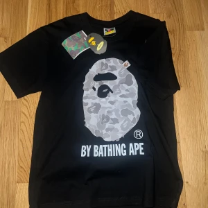 BAPE By Bathing Ape T-shirt Svart - Cool svart T-shirt från BAPE (A Bathing Ape) med klassiskt camo-apehead tryck på bröstet och “By Bathing Ape”  Färg: Svart med grått camotryck passar som M/S