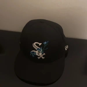 Svart White Sox keps New Era 59FIFTY - Svart keps från New Era 59FIFTY med White Sox-logga framtill och broderade blå blommor. Sidan har World Series-märke i blått och vitt. Platt skärm och klassisk basebollstil. Insidan har 59FIFTY-band och MLB-sticker under skärmen.