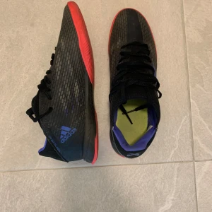 Adidas svarta fotbollsskor med röd sula - Säljer ett par svarta Adidas fotbollsskor med röd sula och blå detaljer. Skorna har snörning och är tillverkade i syntetmaterial. Perfekta för dig som vill ha stilrena och sportiga skor på planen.