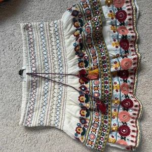Broderad blommig kortkjol med volang - Superfin kortkjol i vitt med färgglada broderade blommor och mönster i rött, blått, gult och grönt. Kjolen har volangkant, snörning med tofsar i midjan och är gjord i bomull. Perfekt för dig som gillar boho-stil och detaljer.