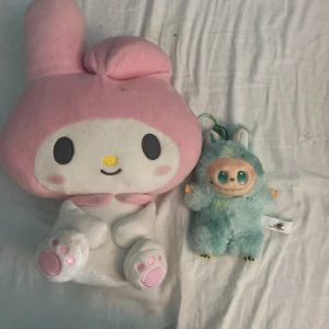 My melodi och lalbubu/lafufu - Bilden visar två gosedjur/leksaker: en stor My Melody-plush från Sanrio och en mindre blå plyschfigur med gröna ögon och kaninöron. Båda ser mjuka ut och är i begagnat skick, men inga tydliga elektronikprodukter syns.