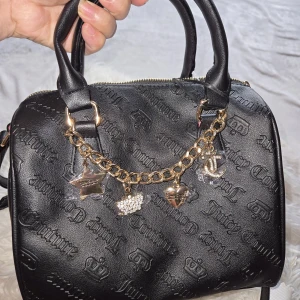 Juicy Couture svart handväska med charms - Svart handväska från Juicy Couture i skinnimitation med präglad logga och kronor över hela väskan. Väskan har två handtag, guldfärgad kedja med söta berlocker och dragkedja upptill. Insidan är fodrad med mönstrat tyg med Juicy Couture-tryck.