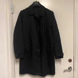 Svart rock från Tiger of Sweden - Stilren svart rock från Tiger of Sweden med klassisk krage och knäppning framtill. Jackan har långa ärmar, två fickor och en rak passform som ger en clean look. Perfekt för dig som gillar minimalistisk stil.