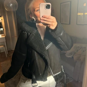 Svart jacka med teddy - Säljer en svart teddyjacka med grov krage och detaljer i skinnimitation. Jackan har bälte nedtill, synliga sömmar och dragkedjor framtill. Fodret och kragen är fluffigt teddy-material som ger en cool look. Perfekt för dig som gillar streetstyle och vill ha något varmt.