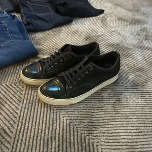 Svarta sneakers från Lanvin - Snygga svarta sneakers från Lanvin med vit sula och glansig tå i skinn. Klassisk låg modell med snörning och diskreta sömmar. Perfekta för dig som gillar stilrena och exklusiva skor med en modern touch.