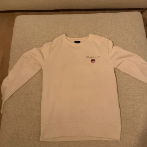 Vit sweatshirt från GANT - Snygg vit sweatshirt från GANT med broderad logga och sköld på bröstet. Tröjan har rund halsringning, långa ärmar och ribbade muddar vid ärmslut och nederkant. Perfekt för en clean och stilren look.