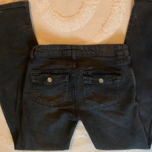 Svarta jeans med ficklock, Perfect Jeans - Köpt för 500kr. Svarta jeans från Perfect Jeans med raka ben och två stora fickor med lock och silverknappar bak. Klassisk femficksmodell med bälteshällor och knappstängning. Snygg, enkel design i kraftigt jeanstyg som passar till det mesta.