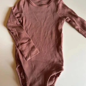 Rosa ribbad body från Lindex - Supersöt långärmad body från Lindex i en dämpad rosa nyans. Tillverkad i mjuk ribbad bomull som är skön mot huden. Perfekt för både lek och chill. Smidig att ta på och av med tryckknappar vid axeln.
