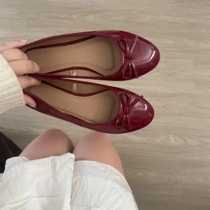 Vinröda ballerinaskor med rosett - Snygga vinröda ballerinaskor i glansigt syntetmaterial med rund tå och söt rosettdetalj framtill. Skorna har platt sula och klassisk design som passar perfekt till både kjol och jeans. Superfina att styla till olika outfits för en chic look.