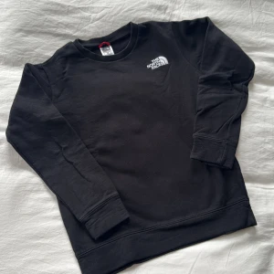 Svart sweatshirt från The North Face - Svart sweatshirt från The North Face med logga broderad på bröstet och tryckt på ryggen. Klassisk rund halsringning, ribbade muddar och mjukt bomullsmaterial. Perfekt för chill dagar eller när du vill ha en clean streetstyle.