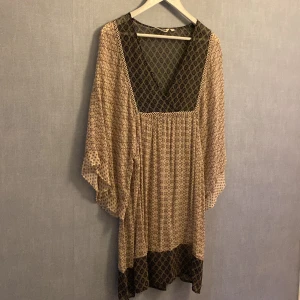 Mönstrad kortklänning från Indiska - Boho-inspirerad tunika från Indiska med v-ringning och vida ärmar. Klänningen har ett unikt mönster i beige och svart . Tillverkad i ett lätt, luftigt material som ger en flowy känsla. Perfekt för dig som gillar avslappnad och trendig stil.