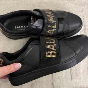 Svarta Balmain sneakers med gulddetaljer - Snygga svarta sneakers från Balmain med bred resår över foten dekorerad med guldfärgade nitar och logga. Skorna är i skinn och har platt sula samt gulddetaljer bak på hälen. Längd på innersulan är 27 cm. Använda 1 gång men aldrig kommit till användning efter det tyvärr. Lite större i storleken!