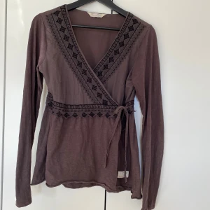 Brun omlott tröja från Odd Molly - Supersnygg brun omlott tröja från Odd Molly med långa ärmar och broderade detaljer längs v-ringningen och midjan. Toppen har knytband i sidan och ett litet broderat motiv på ryggen.