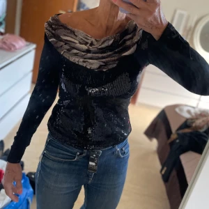 Svart paljettopp med mönstrad krage - Snygg svart topp från Bevell med långa ärmar och glittriga paljetter framtill. Toppen har en bred, draperad krage i grått och svart mönster som ger en cool kontrast. Perfekt för dig som vill sticka ut med en unik och trendig look.