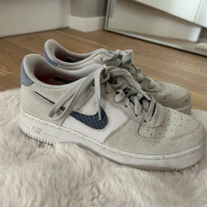 Nike Air Force 1 i ljusgrå mocka - Nike Air Force 1 sneakers i ljusgrå mocka med blå detaljer och vit sula. Swoosh-loggan har ett rutigt blått mönster och insidan är fodrad med röd textil. Klassisk rund tå, snörning och perforeringar på tån för extra ventilation. Perfekta för dig som gillar stilrena och sportiga sneakers.