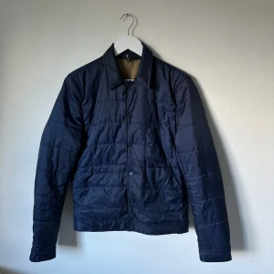 Marinblå quiltad jacka J.Lindeberg S - Snygg marinblå quiltad jacka från J.Lindeberg i storlek S. Jackan har klassisk krage, knäppning framtill och är tillverkad i 100% polyester. Perfekt för dig som gillar stilren och enkel design med en modern touch. Modell: Delroy 41 QLT Feather Nylon. Q