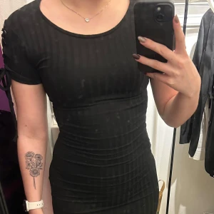 Svart ribbad klänning med snörning på axlarna - Svart ribbad klänning från Atmosphere med tight passform. Klänningen har korta ärmar med cool justerbar snörningsdetalj och rund halsringning. Materialet är en mix av polyester, viskos och elastan som ger stretch och skön känsla. Går precis ovanför knäna på mig.