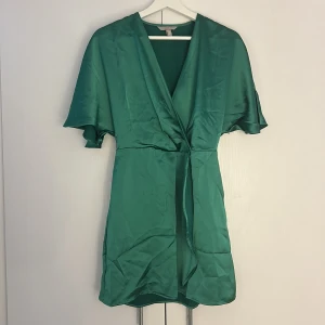 Grön omlottklänning från H&M strl S - (Dragkedjan var sönder när jag köpte den. Därav har jag sytt ett stopp så att den endast går att öppna halvvägs. Syns inget på utsidan och stör inte funktionen.) Snygg grön kort omlottklänning från H&M i glansig polyester. Klänningen har v-ringning, vida korta ärmar och knyt i midjan. Dold dragkedja i sidan för enkel påklädning. Perfekt för dig som vill sticka ut med en färgstark look. Endast använd vid ett tillfälle.