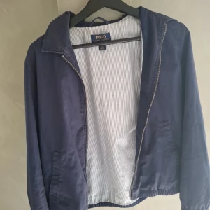 Blå vindjacka från Polo Ralph Lauren - Snygg marinblå vindjacka från Polo Ralph Lauren med klassisk krage, dragkedja och elastiska muddar. Jackan har två fickor framtill och diskret broderad logga på bröstet. Insidan är fodrad med ljusblått och vitt randigt tyg. Perfekt för vår och höst! Storlek 14/16