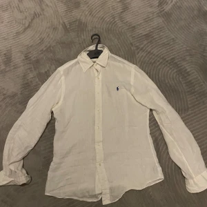 Vit linneskjorta från Ralph Lauren - Klassisk vit skjorta från Ralph Lauren i svalt linne. Skjortan har lång ärm, knappar framtill och den ikoniska blå broderade loggan på bröstet. Perfekt för en clean och stilren look med avslappnad känsla. Den är använd fåtal gånger. Storlek S! OBS. Ett litet hål vid en knapp men som ej är synligt när man har skjortan på sig!