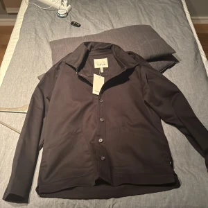 Svart overshirt från !Solid - Stilren svart overshirt från !Solid med klassisk krage och knäppning framtill. Skjortan har två diskreta sidfickor och raka linjer för en clean look. Perfekt att bära öppen eller stängd. Materialet är mjukt och känns bekvämt mot huden.