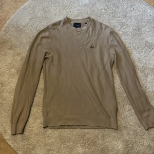 Beige stickad tröja från Lindbergh - Säljer en stilren beige stickad tröja från Lindbergh i storlek M. Tröjan har rund halsringning, diskret broderad logga på bröstet och långa ärmar. Perfekt för lager-på-lager och passar till många olika outfits.