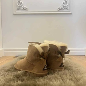 Beiga Everau boots med rosett, stl 38 - Säljer ett par mysiga beiga boots från Everau i storlek 38. Skorna är fodrade med mjuk päls och har en söt rosett bak. Utsidan är i mocka och sulan är mönstrad för bra grepp. Perfekta för kalla dagar när du vill hålla fötterna varma. Aldrig använts. Tyvärr är de för små. Inköpta i Australian.