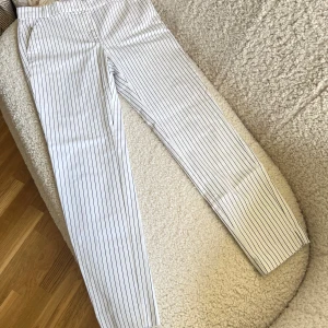 Randiga kostymbyxor från Mango - Snygga vita kostymbyxor från Mango med tunna svarta ränder. Byxorna har raka ben, fickor fram och en dekorativ bakficka. Tillverkade i ett mjukt bomullsmaterial som känns skönt mot huden. Perfekta för en clean och stilren look.