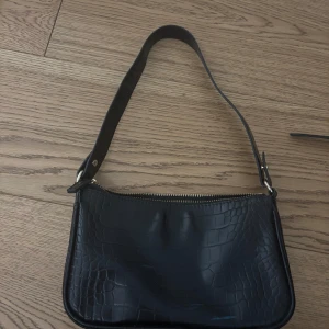 Svart axelväska i croco-imitation - Snygg svart axelväska med croco-mönstrad yta och guldfärgad dragkedja. Väskan har en stilren form och bred axelrem, perfekt för att lyfta vilken outfit som helst. Materialet är syntet med struktur som påminner om krokodilskinn.