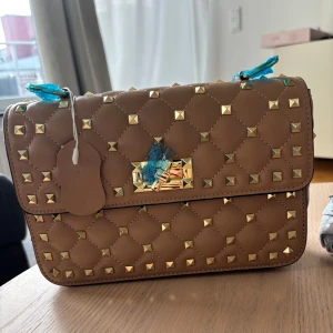 Liknande valentino väska med nitar - Snygg beige axelväska i äkta skinn med guldiga nitar över hela väskan. Väskan har ett vridlås i guld och en kedjerem. Den är inte äkta valentino med väldigt lik. Den har även aldrig använts och är helt ny i bra skick. Tveka inte om att fråga något💗