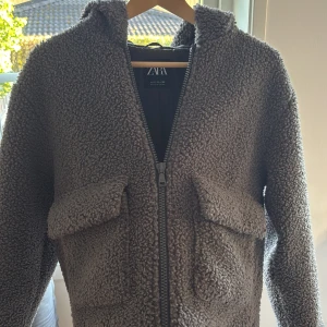 Grå teddyjacka med huva från Zara - Mysig grå teddyjacka från Zara med huva och stora fickor framtill. Jackan har dragkedja och är tillverkad i ett fluffigt teddy-material som håller dig varm. Perfekt för kyliga dagar och enkel att matcha med olika outfits.