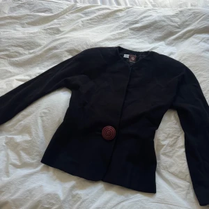 Svart kavaj från Anne Klein med stor knapp - Säljer en svart kavaj från Anne Klein med en unik stor spiralformad brun knapp framtill. Jackan har lång ärm, rund halsringning och är figursydd för en snygg siluett. Perfekt för dig som gillar klassiska plagg med en twist.