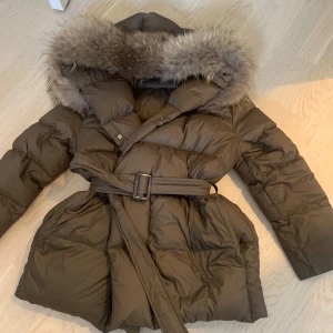 Brun dunjacka med äkta päls och bälte - Snygg brun dunjacka med stor huva kantad av fluffig äkta päls. Jackan har bälte i midjan och långa ärmar, perfekt för kalla dagar. Puffig modell som ger en cool och trendig look.
