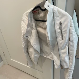 Vit skinnjacka med dragkedjor - Säljer en snygg vit skinnjacka med coola detaljer som dragkedjor och quiltade partier på axlarna. Jackan har en modern bikerstil och är perfekt för dig som vill sticka ut lite extra. Passar till många olika outfits och har lång ärm.